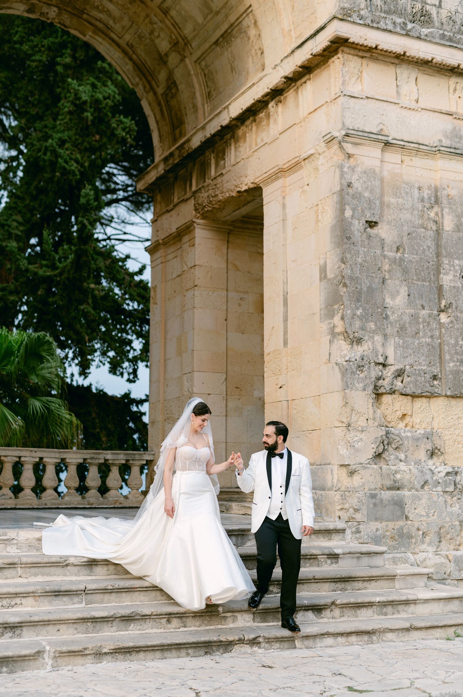 A couple in wedding attire sits and poses on stone steps of a historic building with arched architecture in Corfu Greece