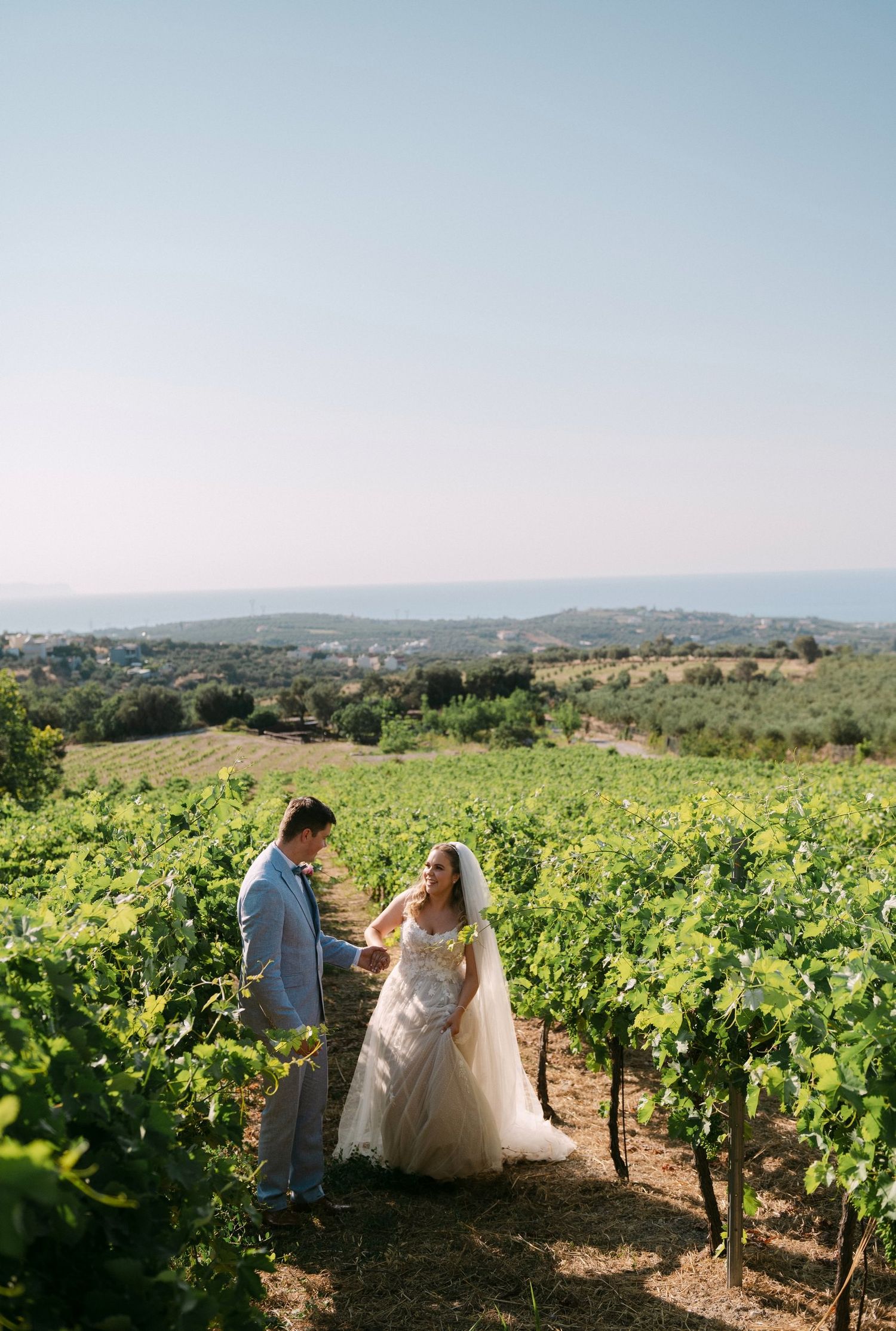 Newlyweds walk hand in hand through scenic vineyard rows during golden hour in wine country.