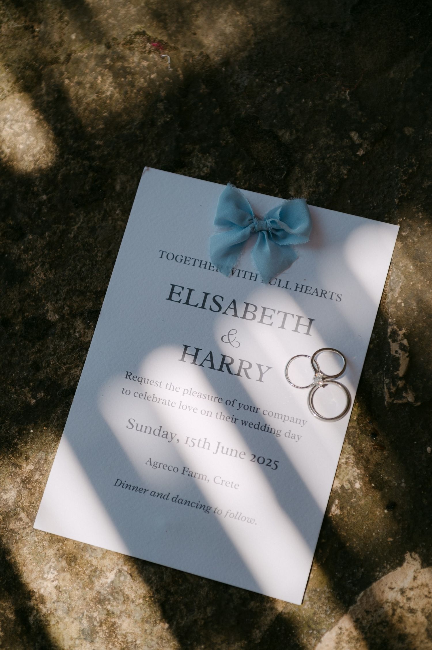 A white wedding invitation card with blue text and ribbon lies in dappled sunlight showing wedding details and interlinked rings.