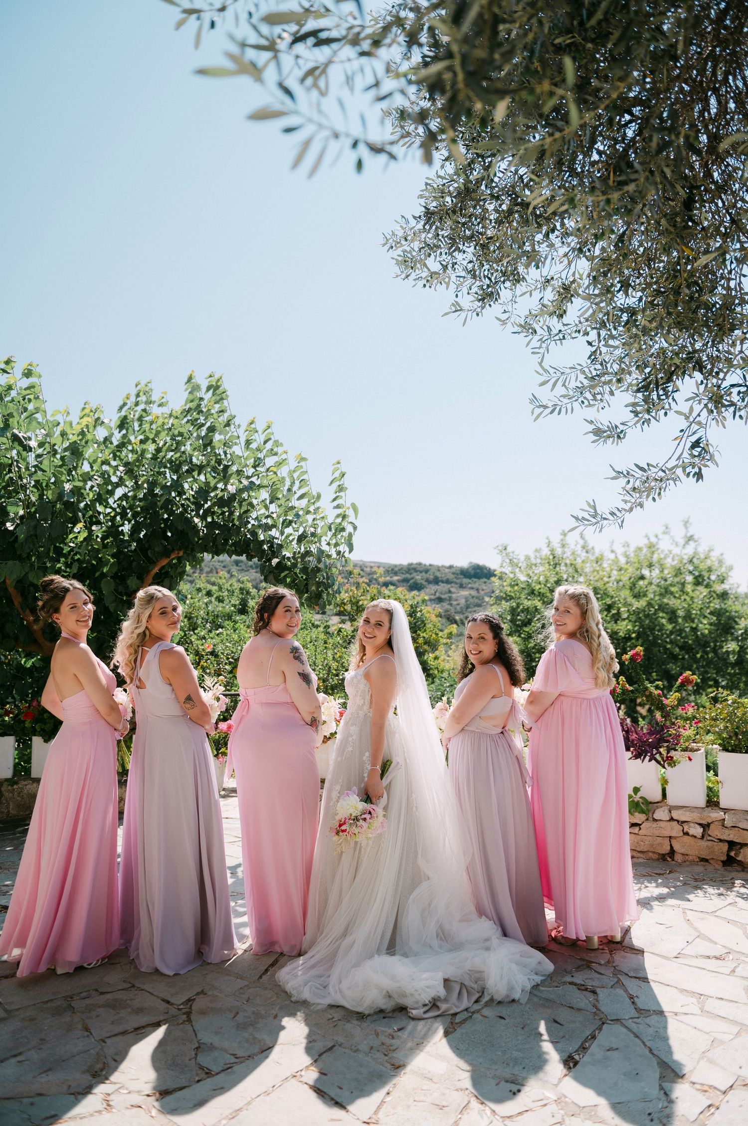 Bridal party in pink and white dresses standing in a line at an outdoor wedding venue on a sunny day.