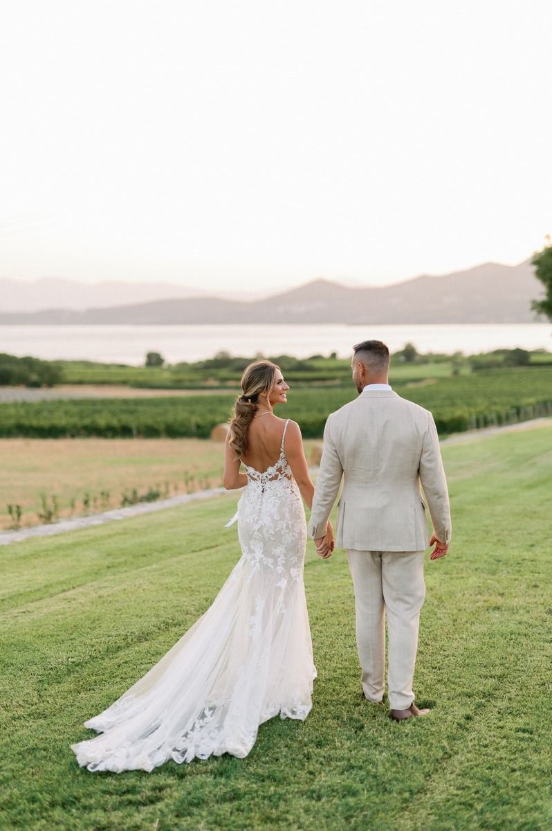 Bride in lace gown and groom in light suit walking on hillside with water view.