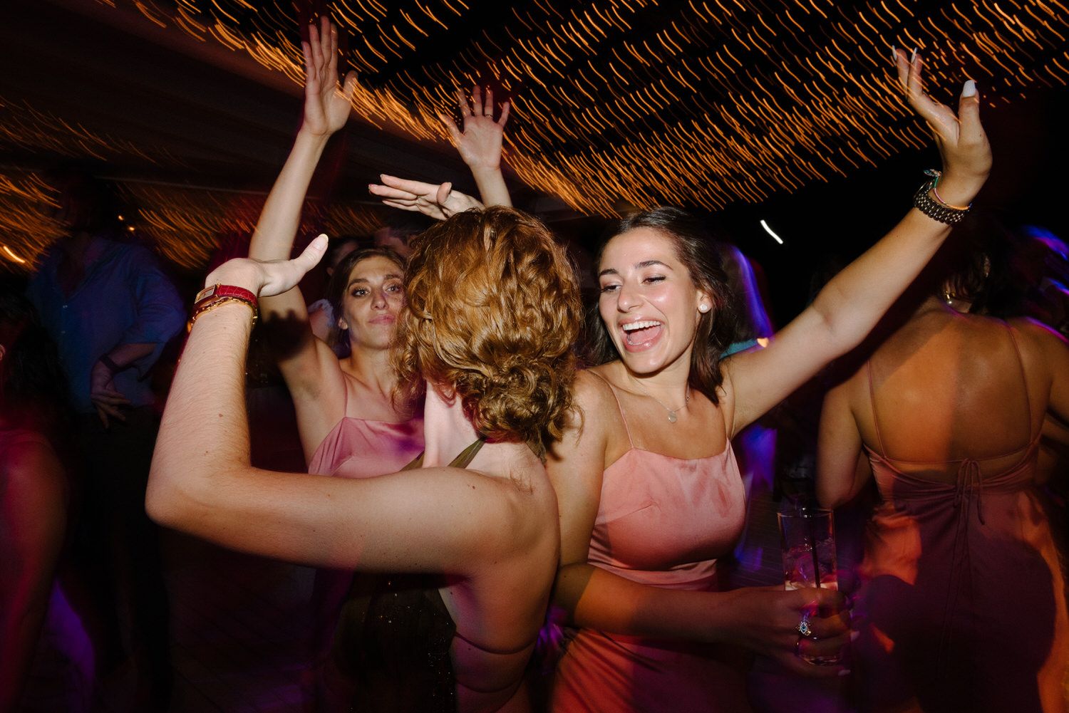 Wedding guests dance with raised arms under twinkling reception lights.
