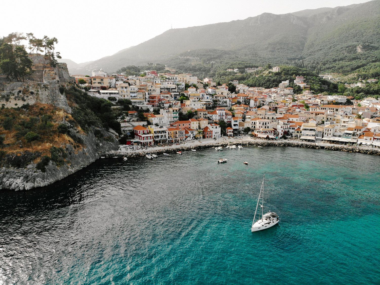 Aerial view of a coastal Mediterranean village with colorful buildings and a sailboat in turquoise waters.