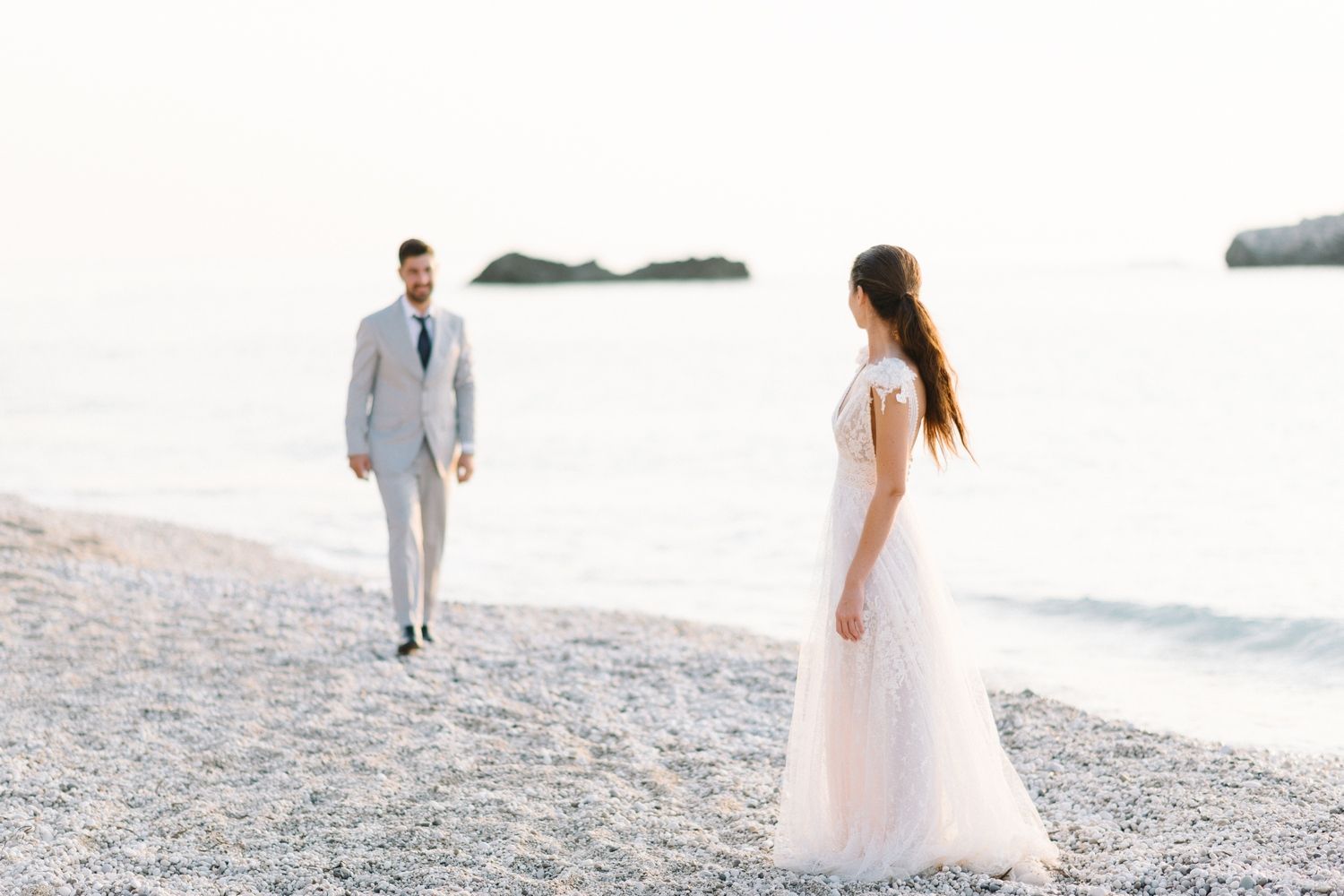 A romantic couple in wedding attire walks along a pebbled beach at sunset with distant islands in view.