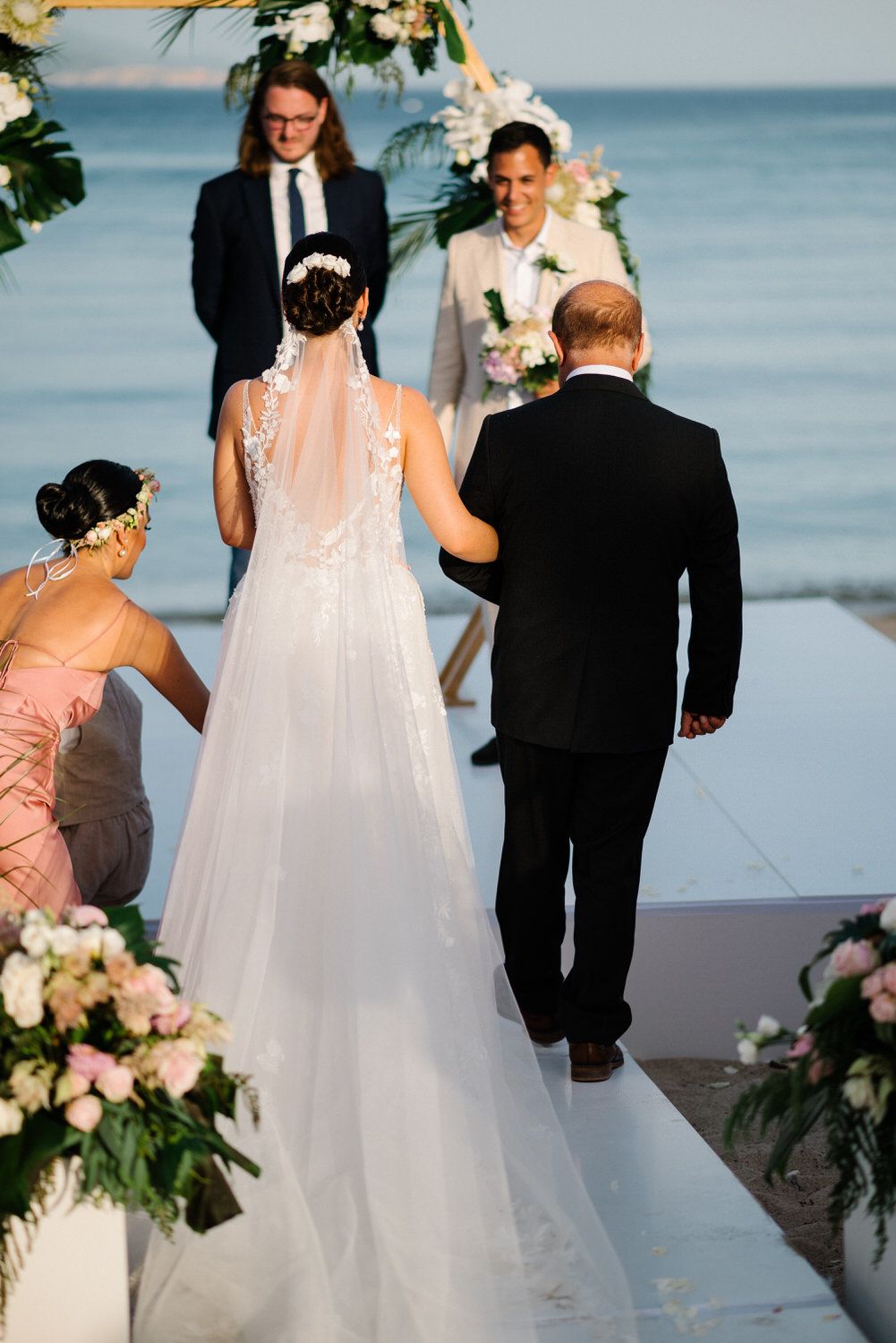 Wedding ceremony on white platform overlooking ocean with floral arch and guests.