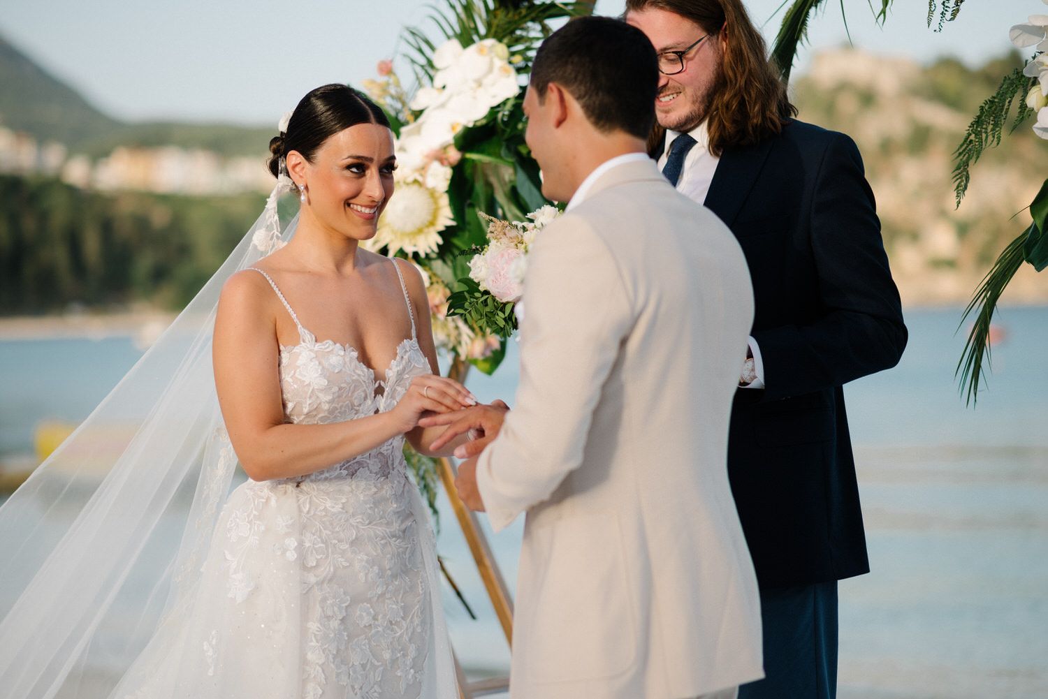 A couple exchanges rings during a beachside wedding ceremony with tropical decor and an officiant.