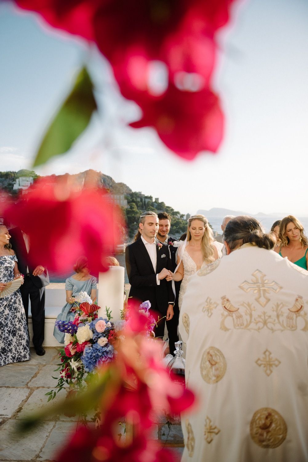A wedding ceremony framed by vibrant red flowers with coastal mountains in the background.