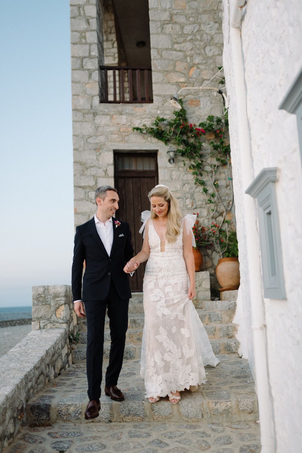 Couple descends stone steps of Mediterranean villa wearing elegant wedding attire.