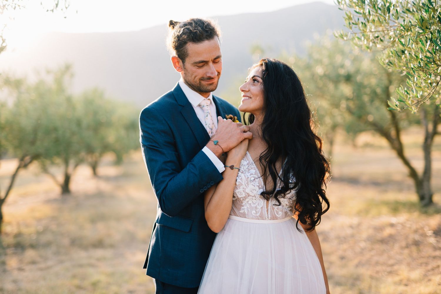 Newlyweds share intimate moment among olive trees at sunset.