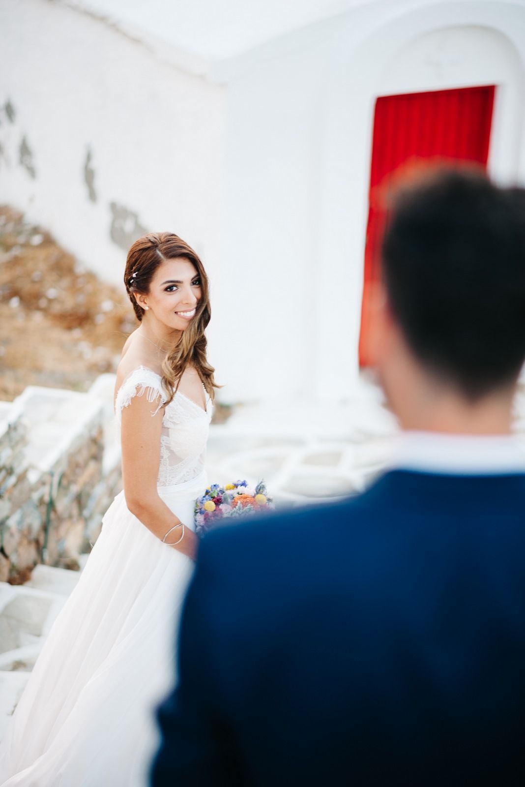 Bride in white dress smiles while holding colorful bouquet near red door.