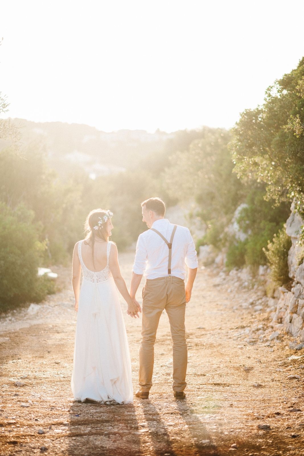 A couple in wedding attire holding hands on a dirt path during golden hour sunset.