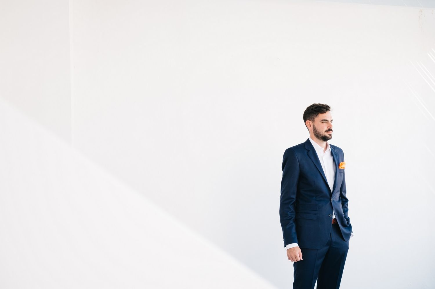 A man in a navy blue suit stands against a white wall in a minimalist setting.