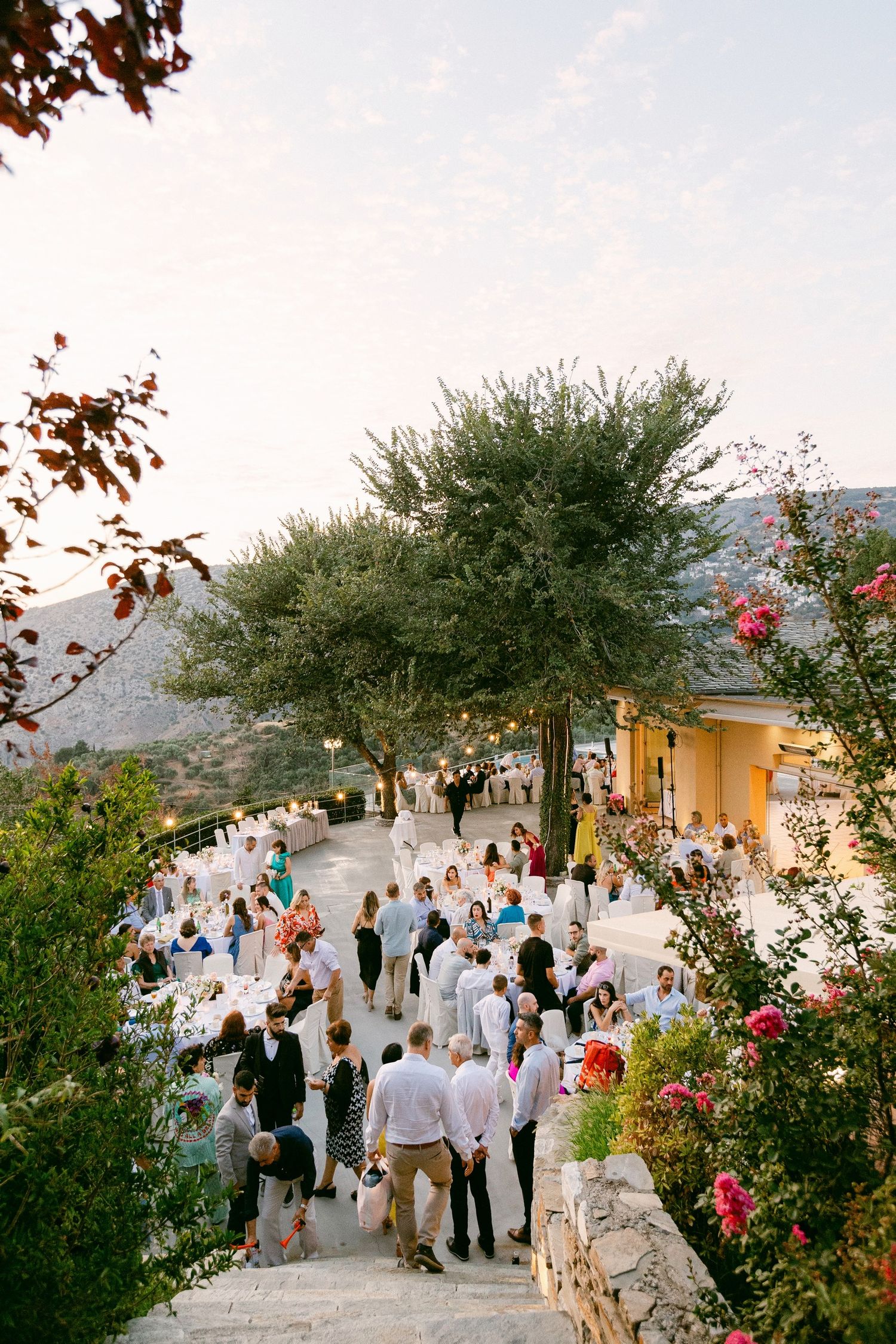 A crowd of guests gather on a winding hillside path surrounded by trees and flowering plants at an outdoor event venue.