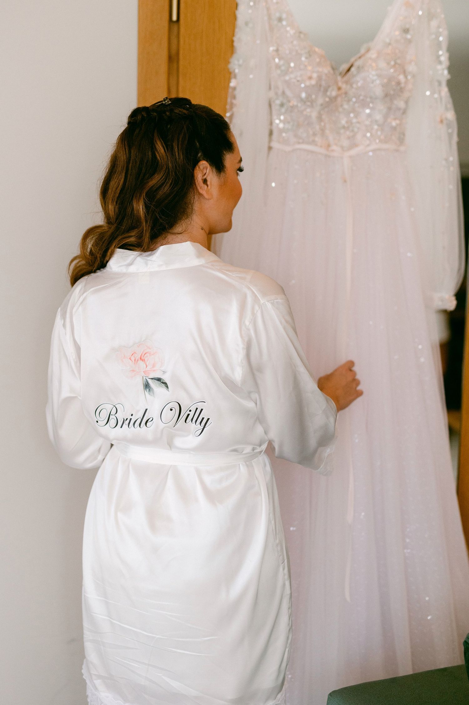 Someone wearing a white silk robe with 'Bride Villa' text embroidered looks at a hanging beaded wedding dress.