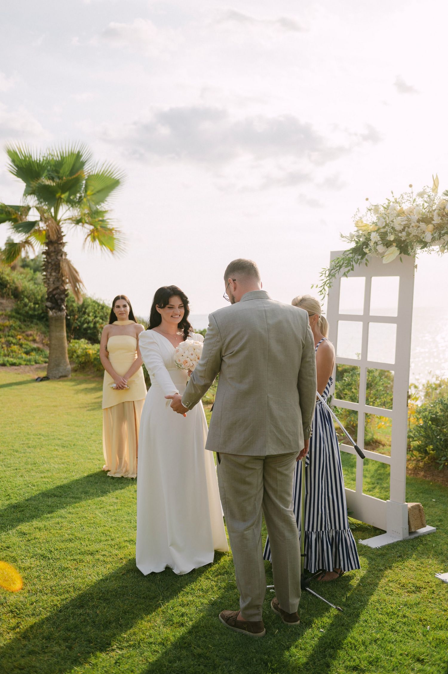 An outdoor wedding ceremony takes place on a grassy lawn with a white arch and palm tree under cloudy skies.