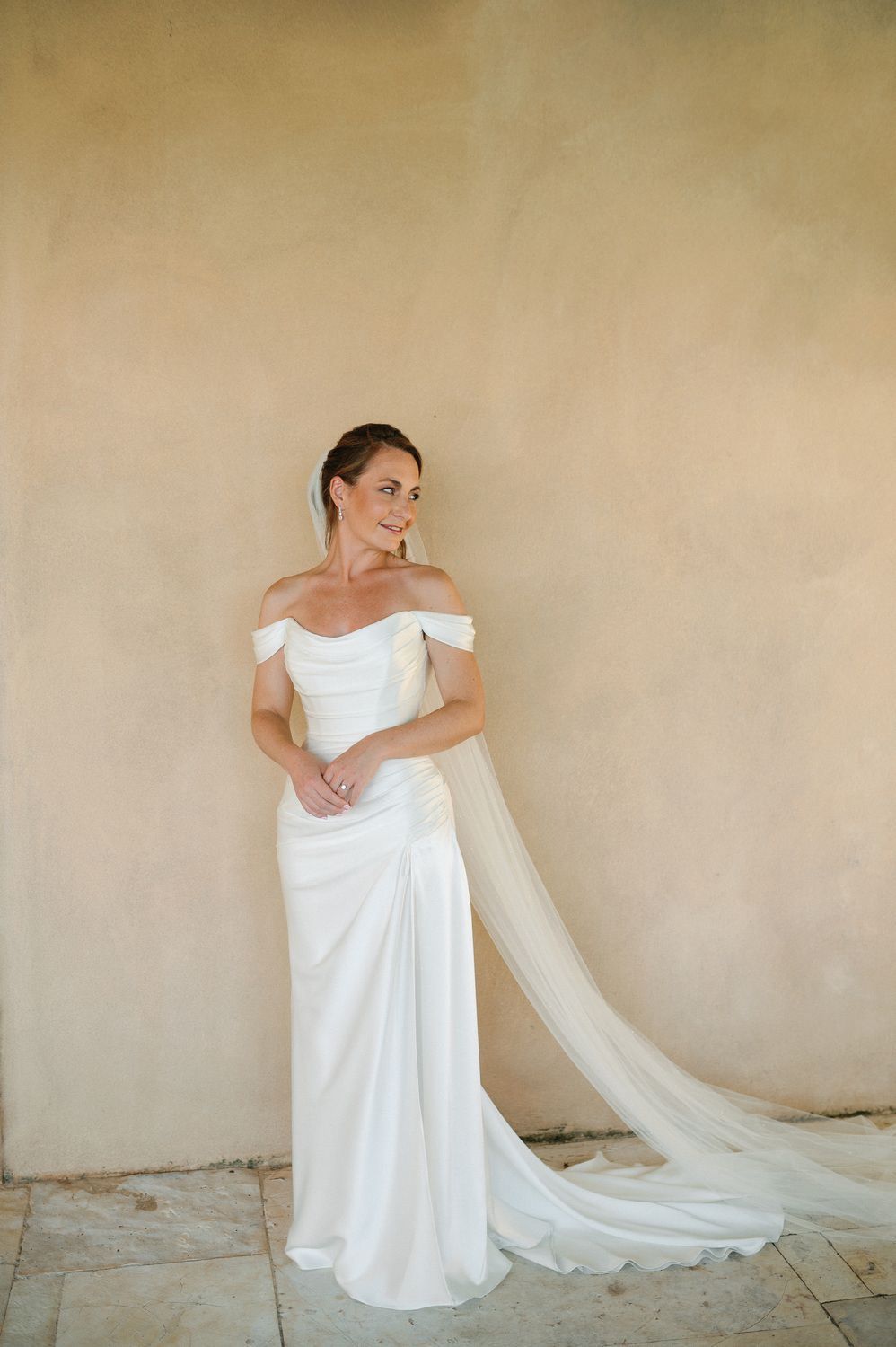 An elegant off-shoulder white wedding gown with a flowing train against a neutral beige background.
