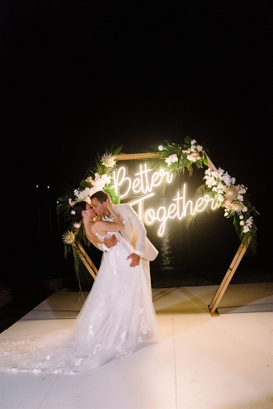 A couple dances romantically under an illuminated 'Together Forever' neon sign decorated with floral arrangements at night.