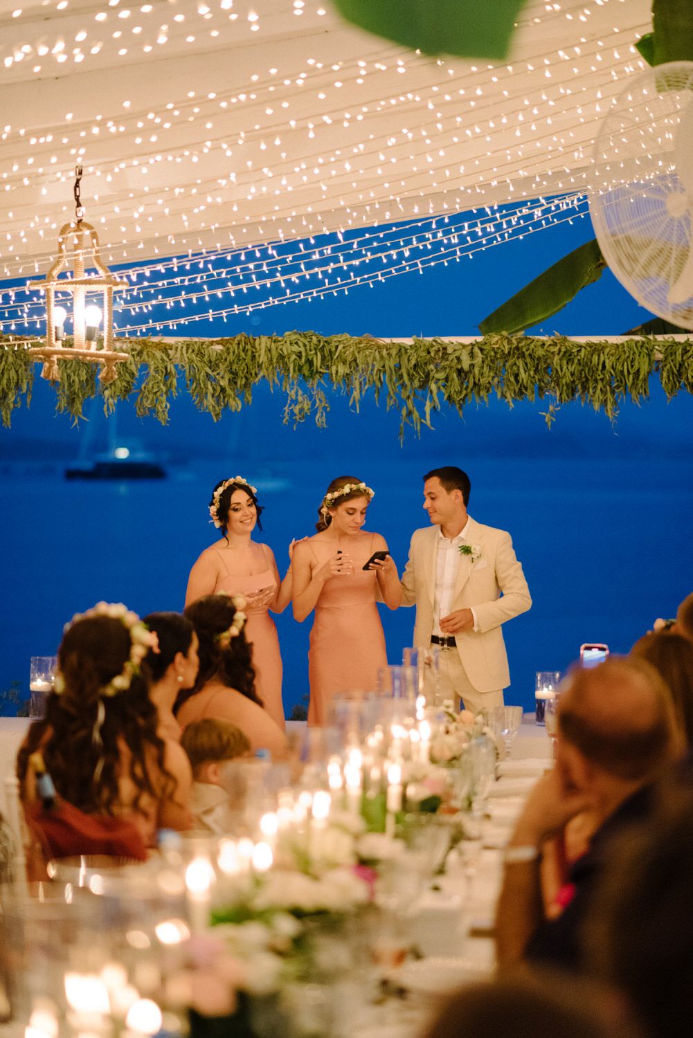 Toast during an evening reception under string lights with ocean backdrop and candlelit tables.