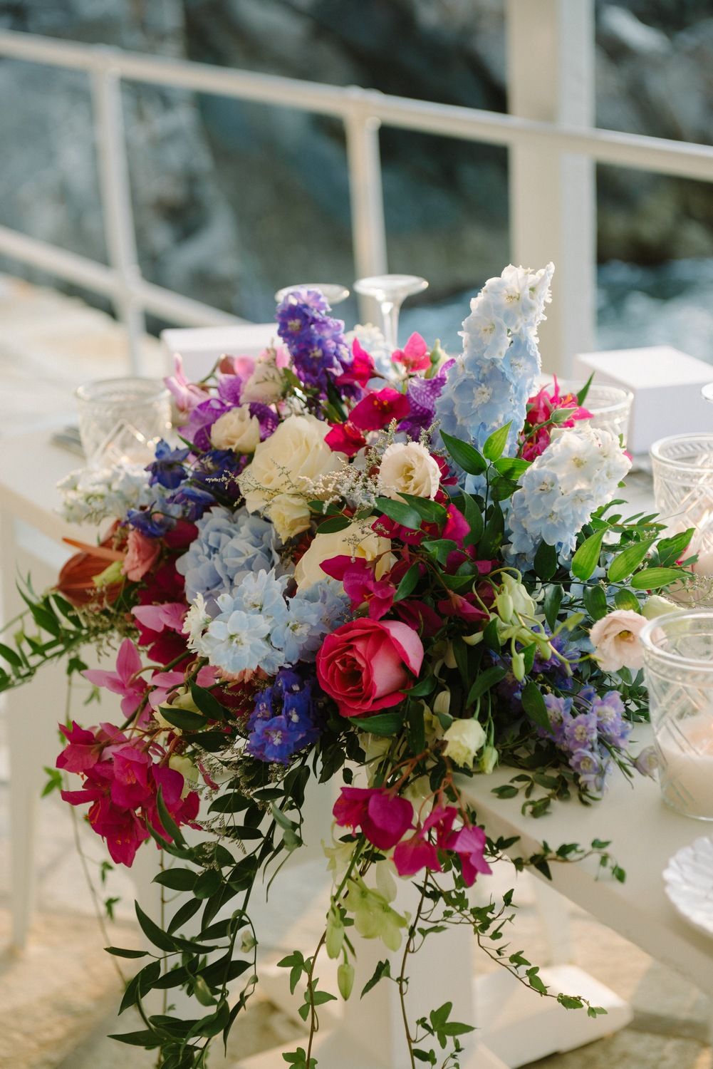 Vibrant floral centerpiece with purple, pink and white flowers arranged with cascading greenery on an elegant table setting.