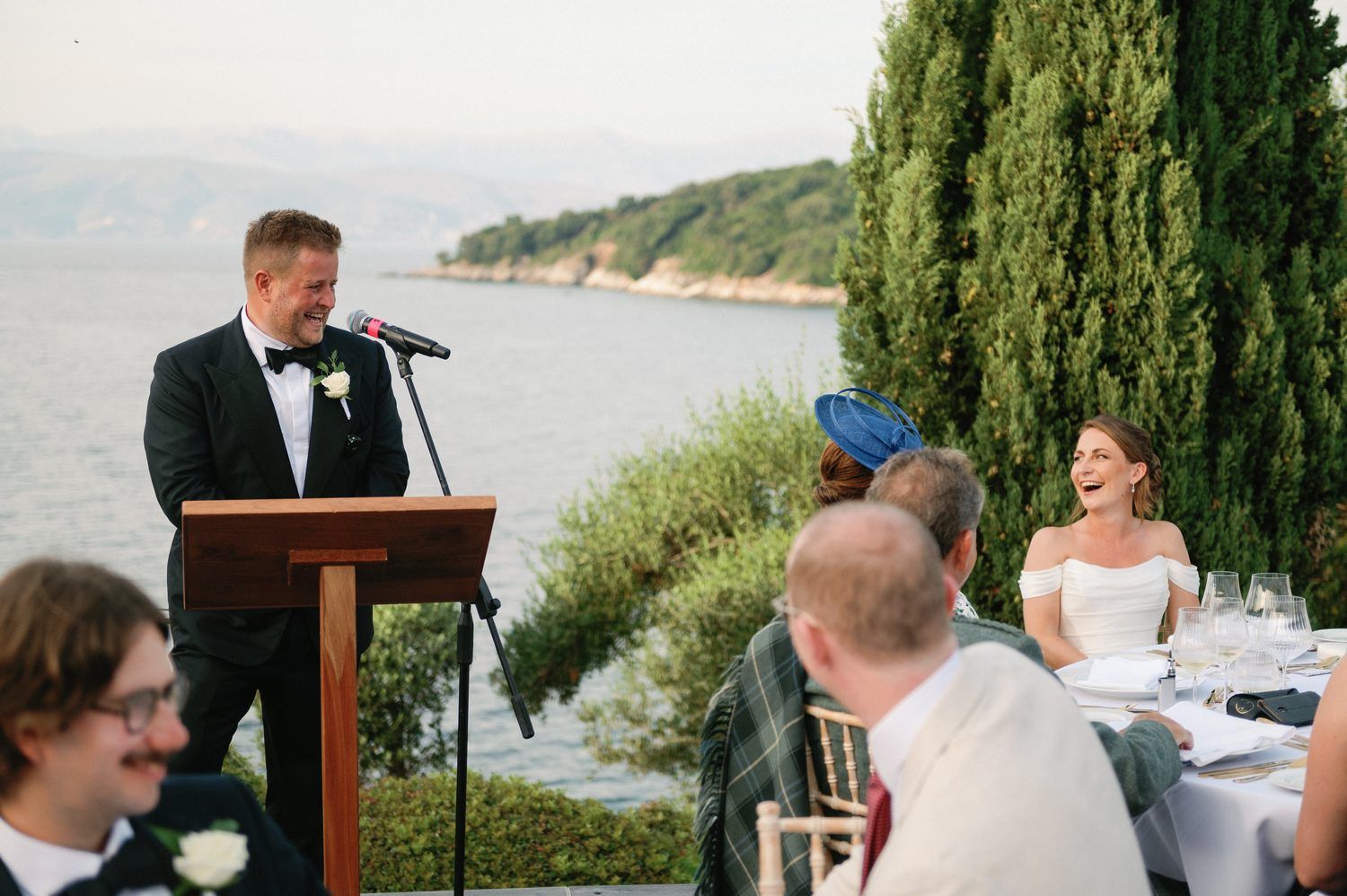 Someone delivers a speech at an outdoor wedding reception podium overlooking a scenic waterfront.