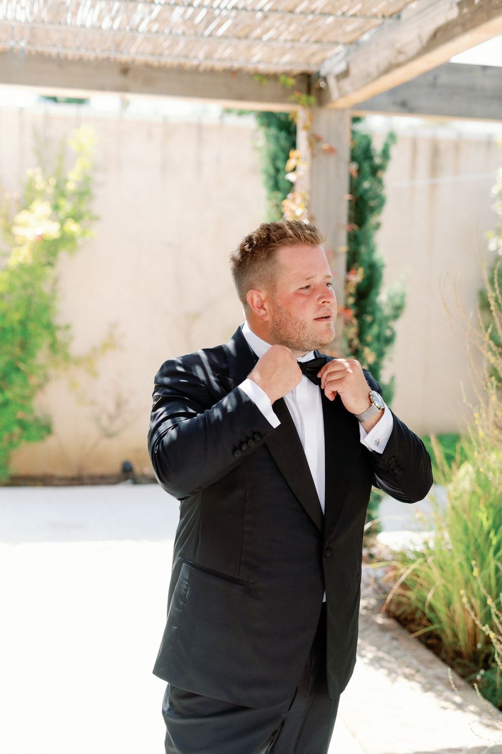 A groom in black tuxedo adjusts his tie while standing under a pergola.