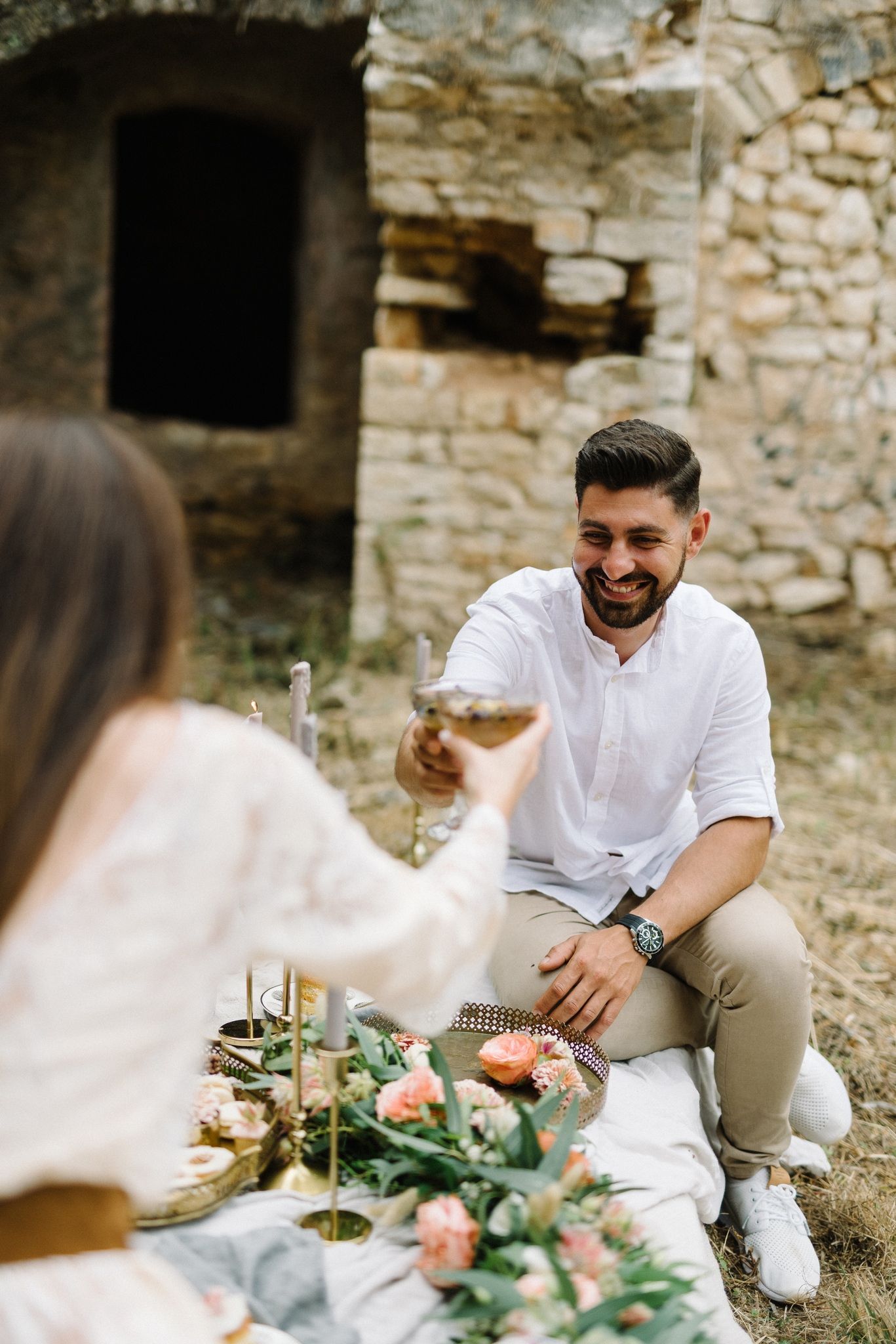 Romantic picnic setup with white-clothed couple toasting wine glasses near stone ruins and floral arrangements.