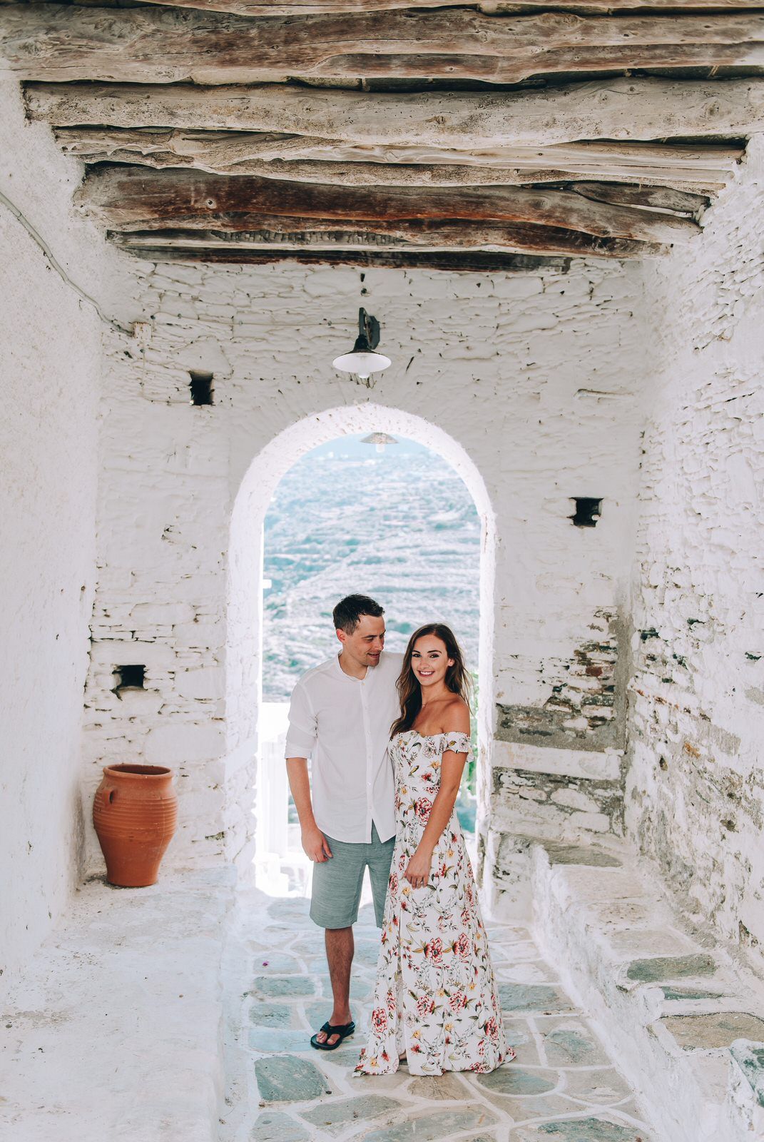 A couple stands in a rustic white archway overlooking the Mediterranean Sea with a terracotta pot nearby.