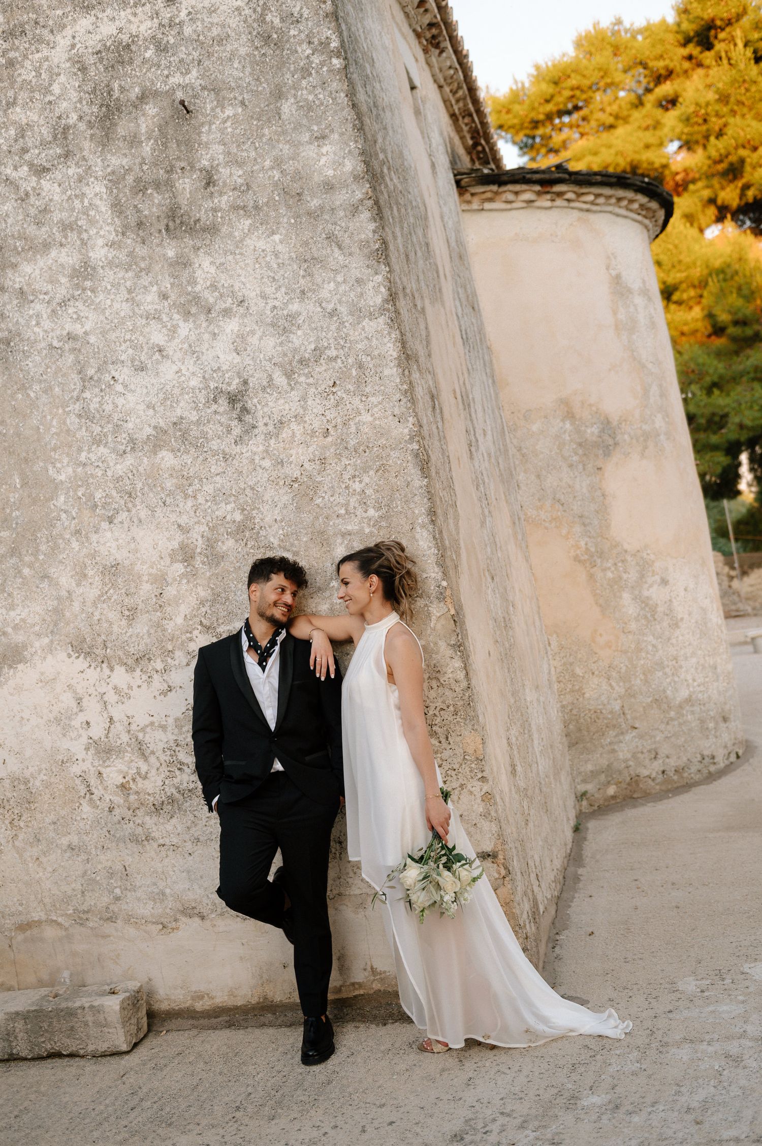 Bride and groom lean against rustic stone wall, she holds white bouquet, autumn trees visible behind them.
