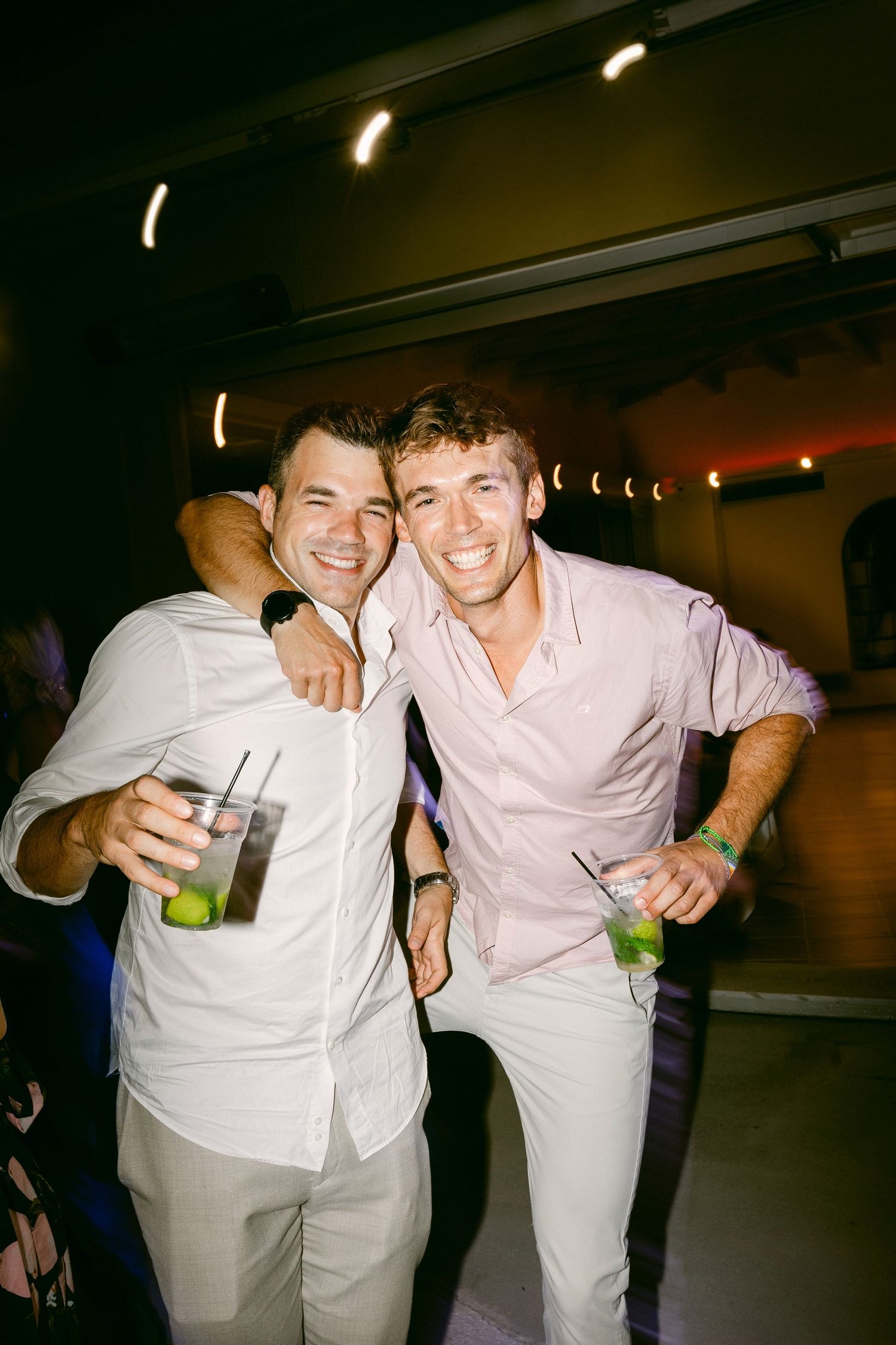 Two friends in light colored clothing smile and pose with drinks at a dimly lit indoor venue during a night out.