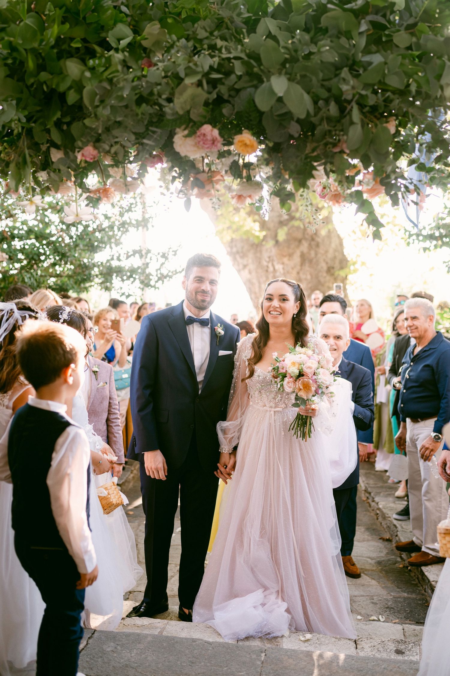 A wedding ceremony takes place outdoors under a floral archway with guests gathered to celebrate the special occasion.