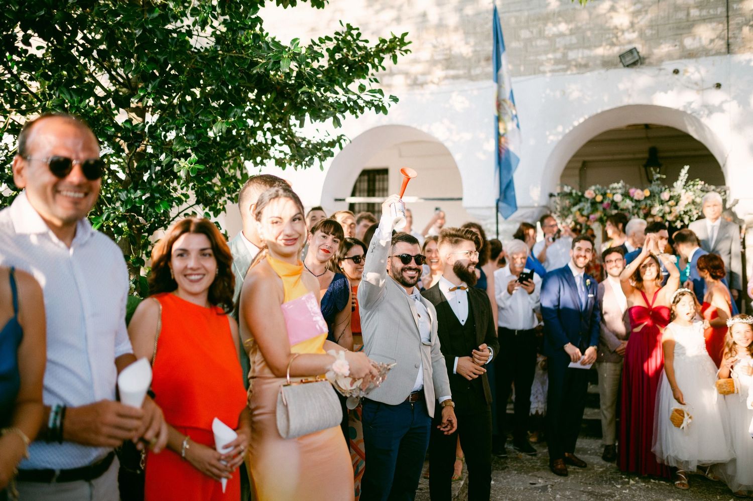 A crowd of wedding guests gather outdoors under stone archways on a sunny day with green foliage overhead.