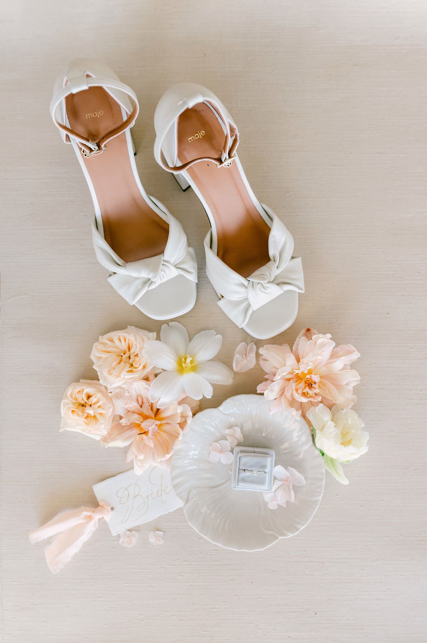 Elegant white bridal shoes with bow details displayed alongside peach garden roses and a perfume bottle on beige surface.