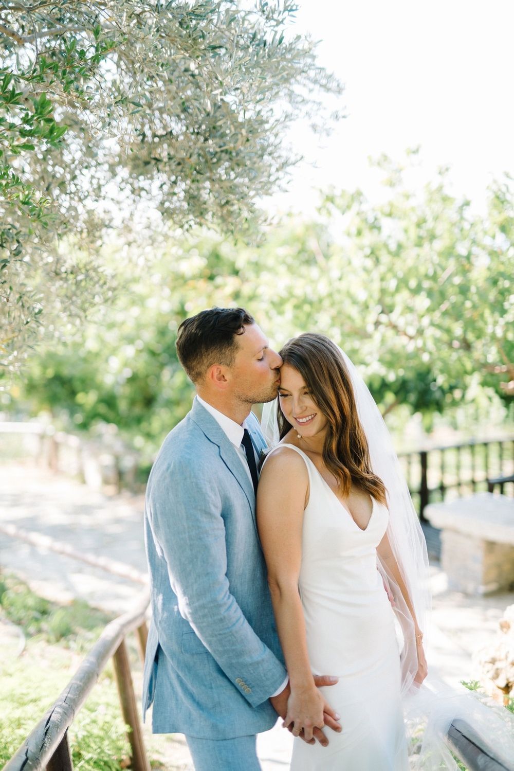 A couple shares romantic moments under blooming trees in a garden setting wearing light blue and white attire.