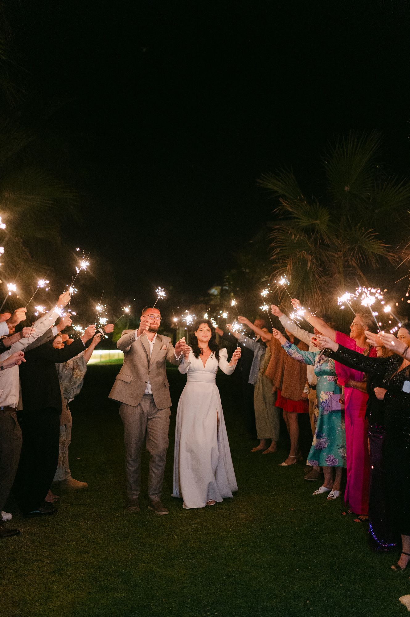 Wedding guests form a sparkler tunnel at night as a couple celebrates their special moment on a lawn with palm trees.