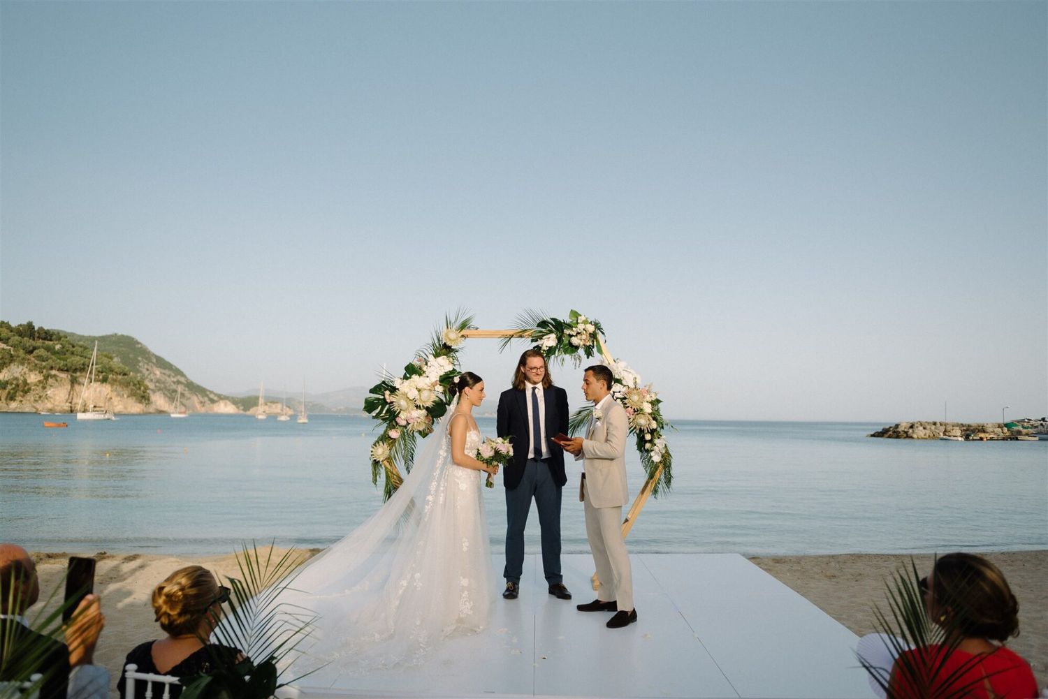 A wedding ceremony takes place on a white platform overlooking a beautiful coastal bay with mountains in the background.
