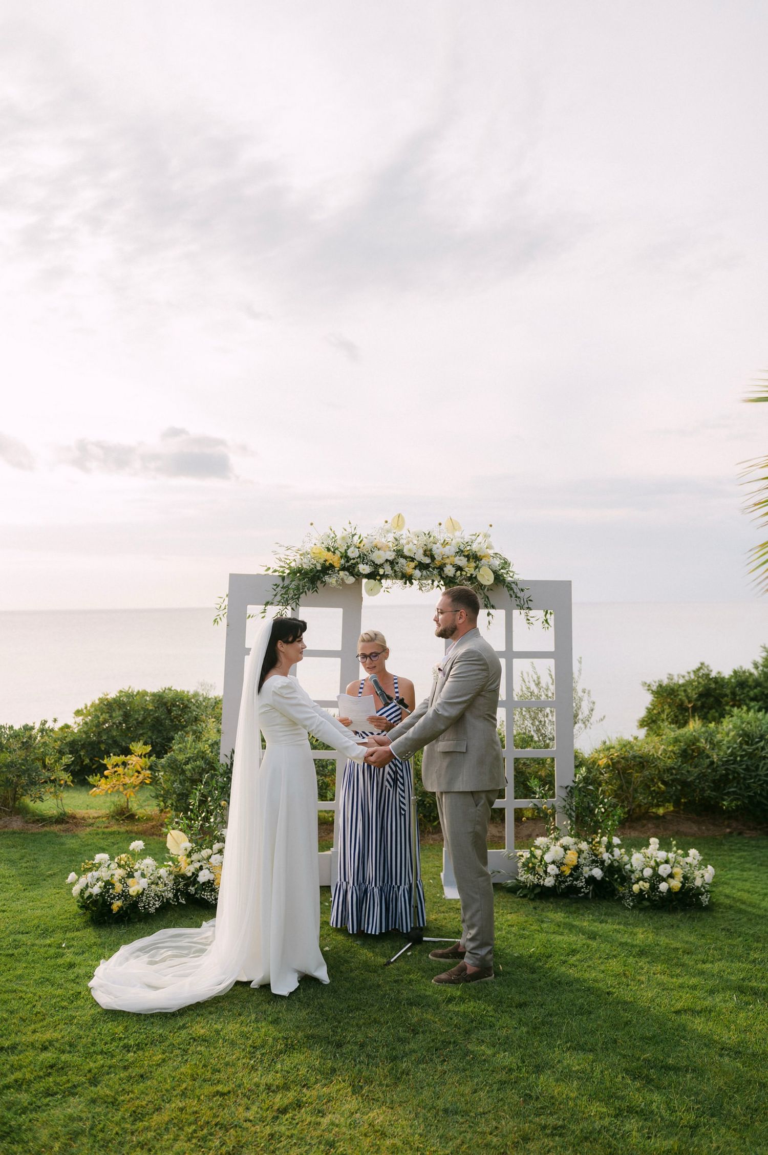 Intimate outdoor wedding ceremony on grassy lawn with white floral arrangements and wooden arch at sunset.