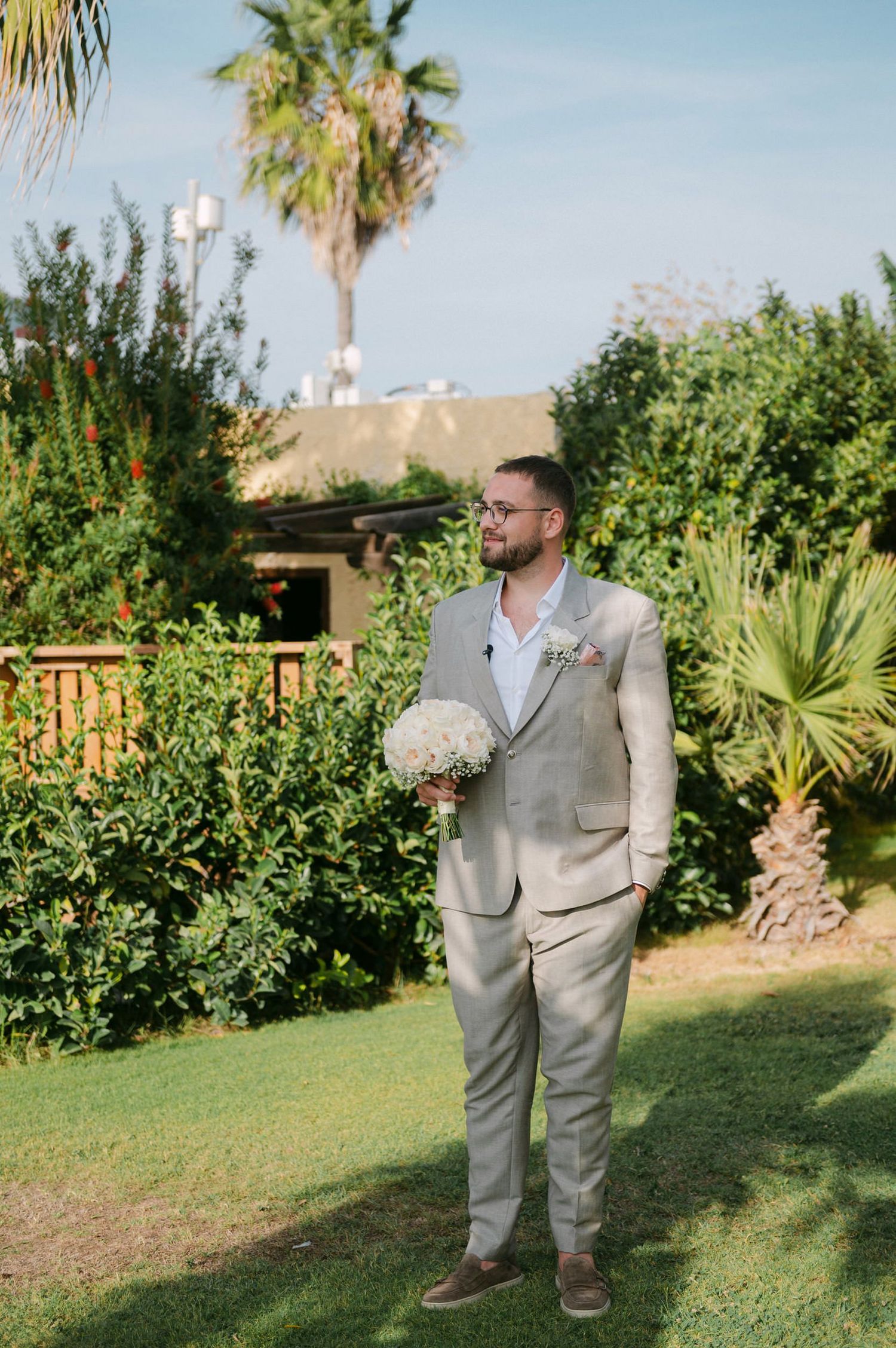 A person in a light gray suit holds a white floral bouquet while standing in a tropical garden setting with palm trees.