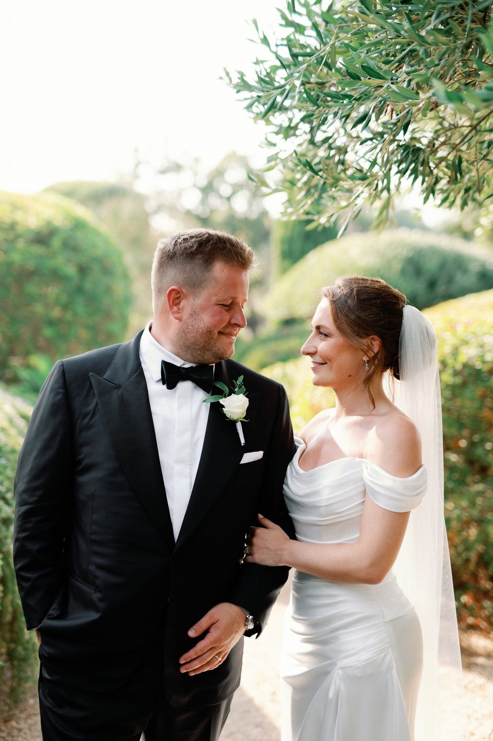 A romantic wedding portrait in a garden with the couple in classic black tuxedo and off-shoulder white gown.