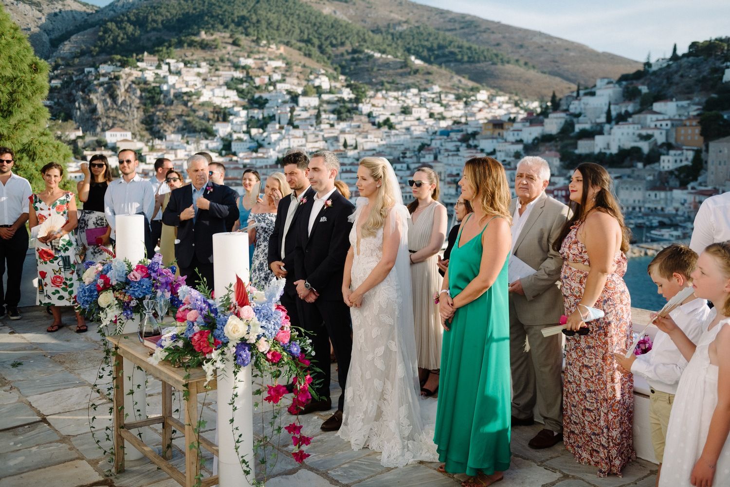 Wedding party and guests gathered on stone terrace with colorful floral arrangements.