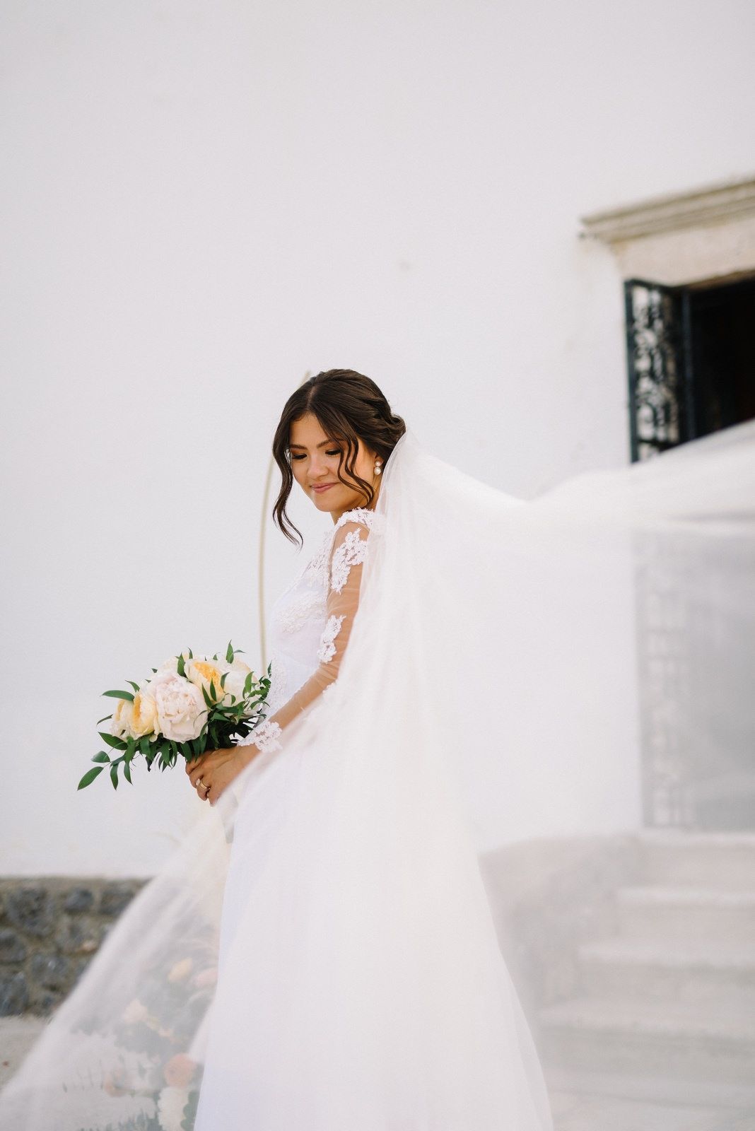 Bride in flowing white dress holding pink bouquet on Mediterranean steps.