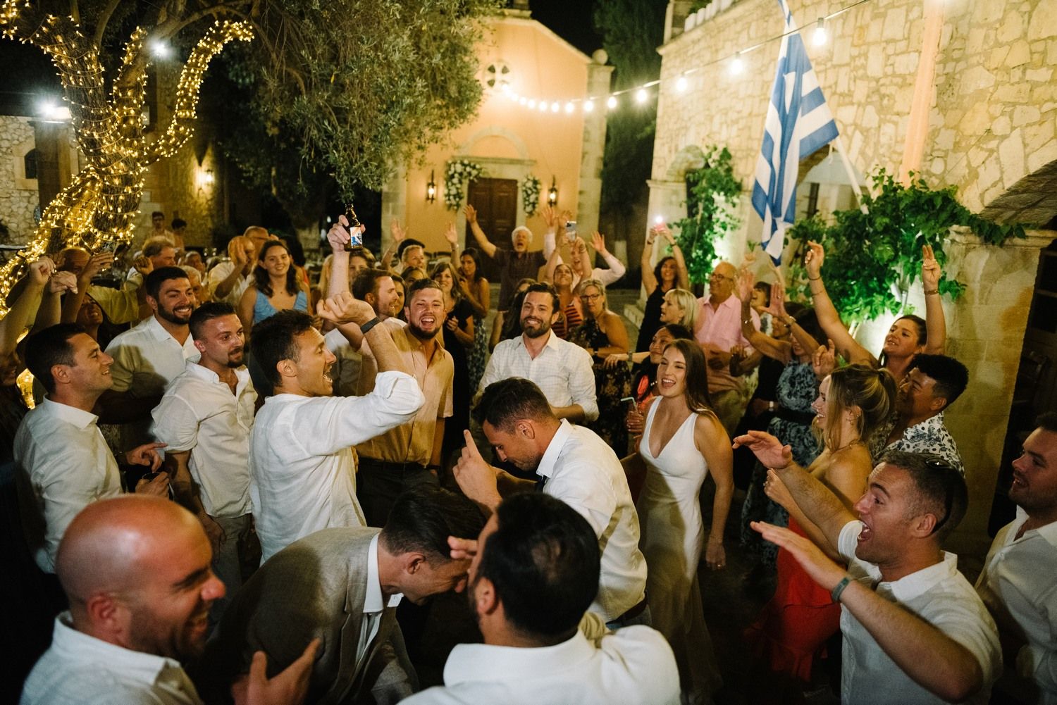 Energetic crowd dancing and celebrating at a nighttime outdoor wedding reception with string lights and stone walls.