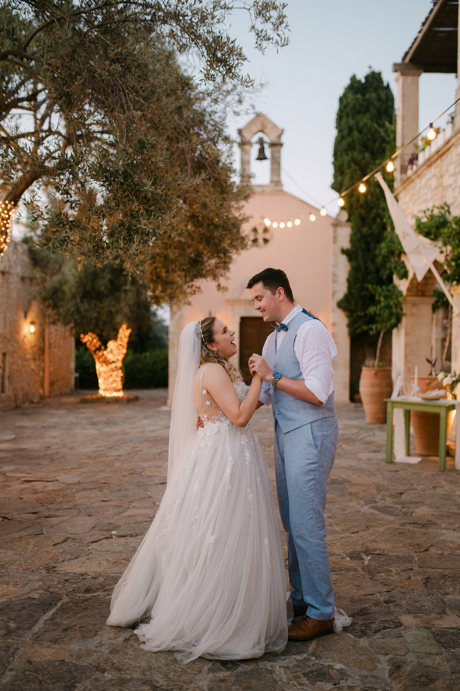 A beautiful Mediterranean wedding scene with a pink chapel and olive trees decorated with twinkling lights.