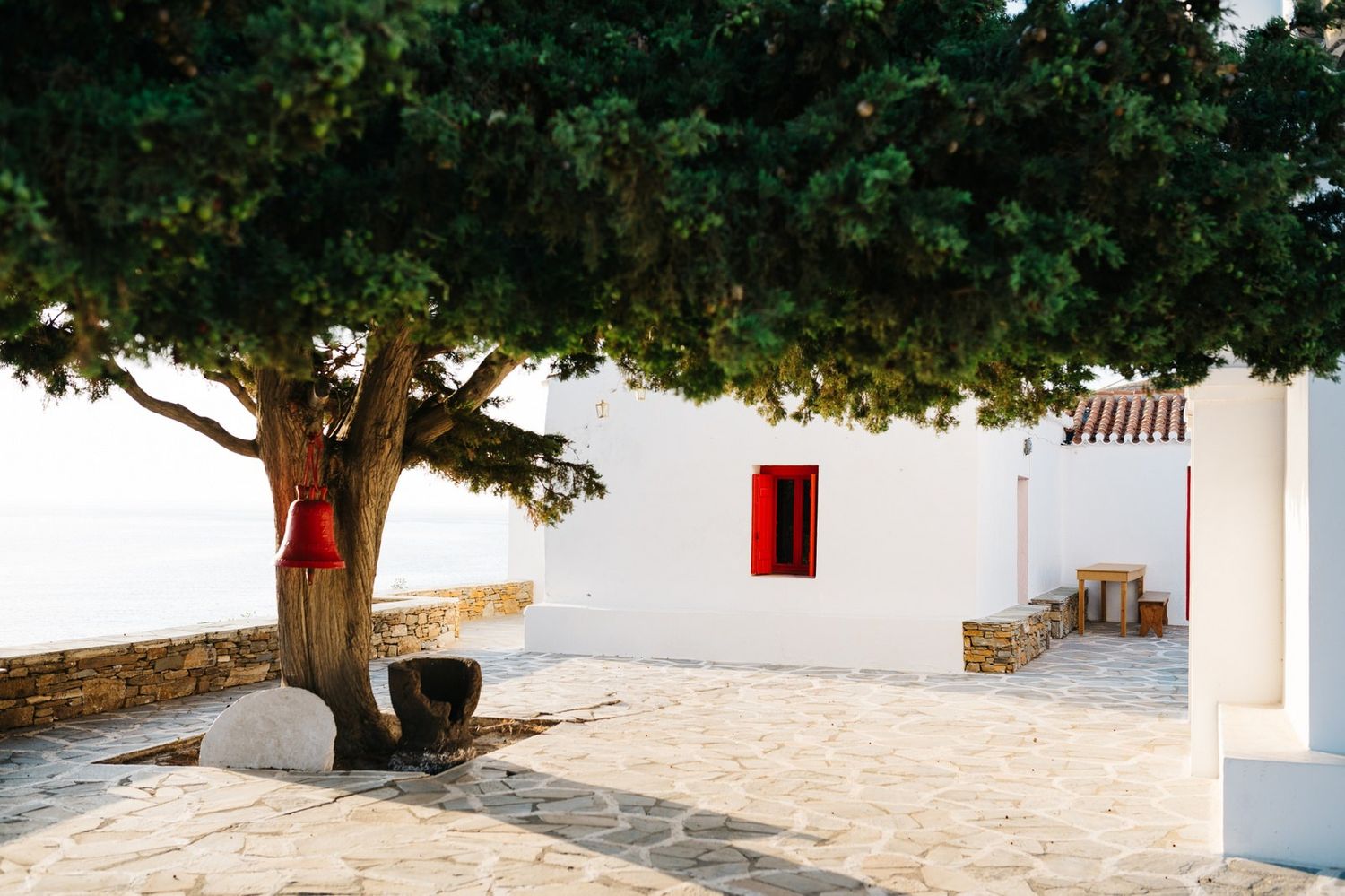 Large tree casting shade over white Mediterranean building with red window frame.