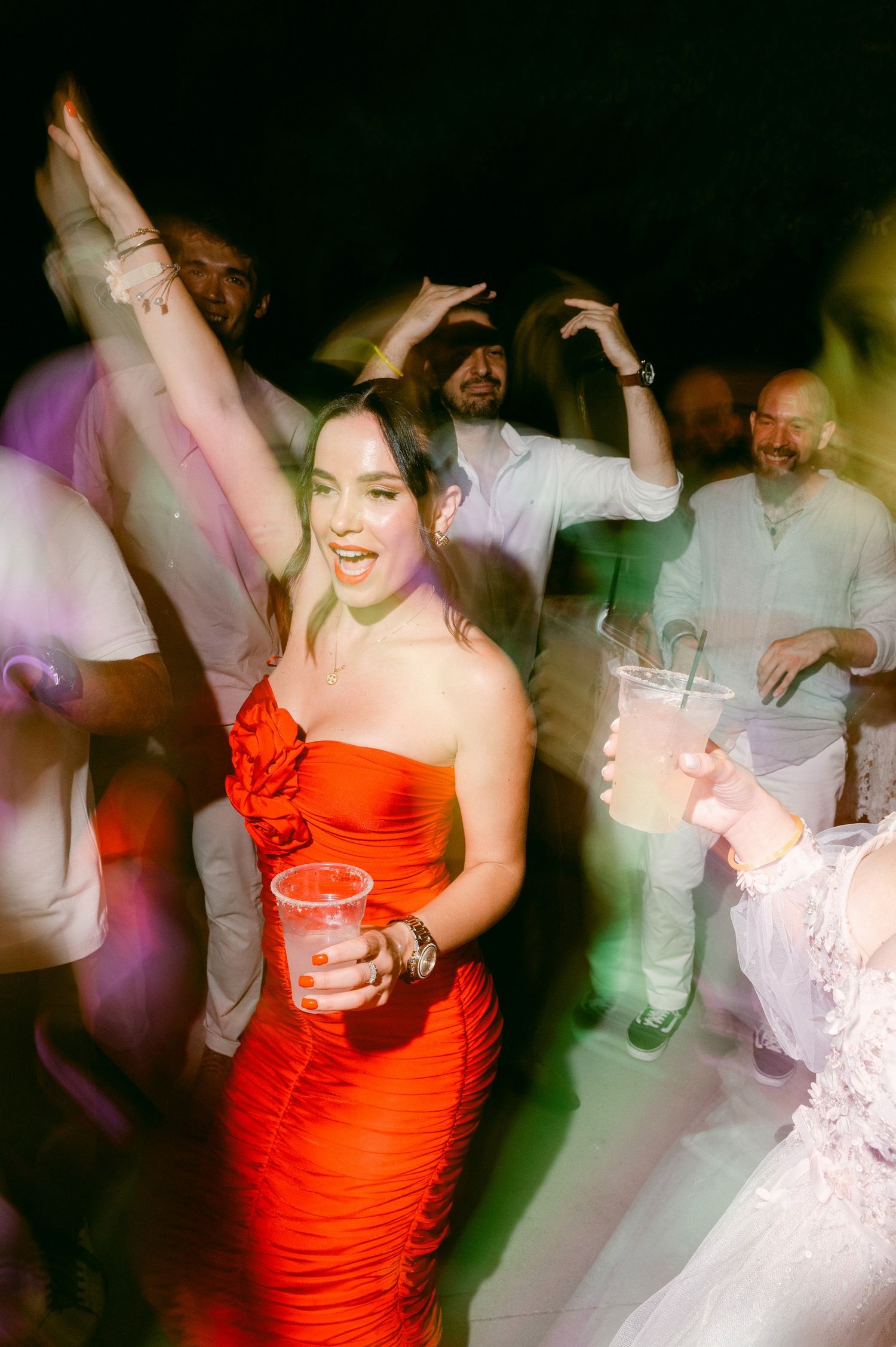 Dancers moving energetically on a dimly lit dance floor with a person in a vibrant red dress holding a drink in the center.