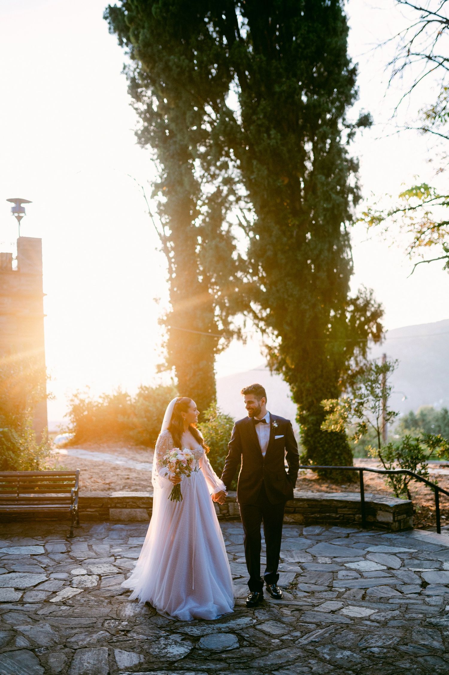 A couple in wedding attire stands together on a stone patio at sunset with tall cypress trees in the background.