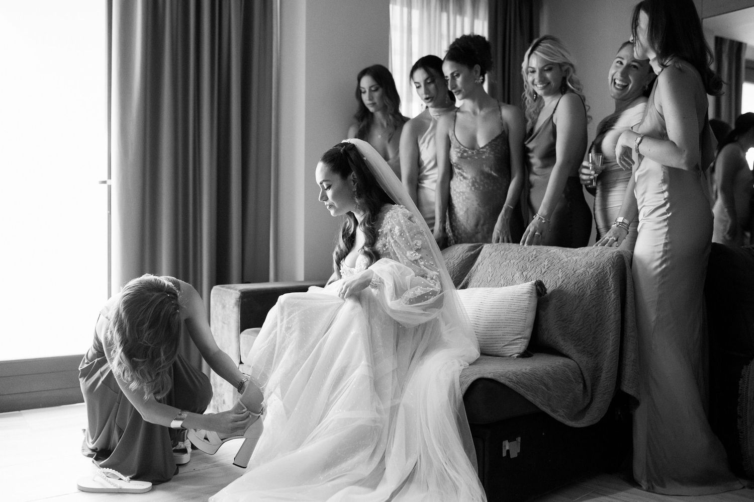 A wedding preparation scene showing people in formal attire gathered around a sofa in a sunlit hotel room.