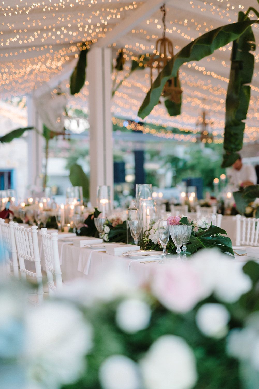 Dreamy wedding reception setup with string lights overhead and tropical leaf table decorations.