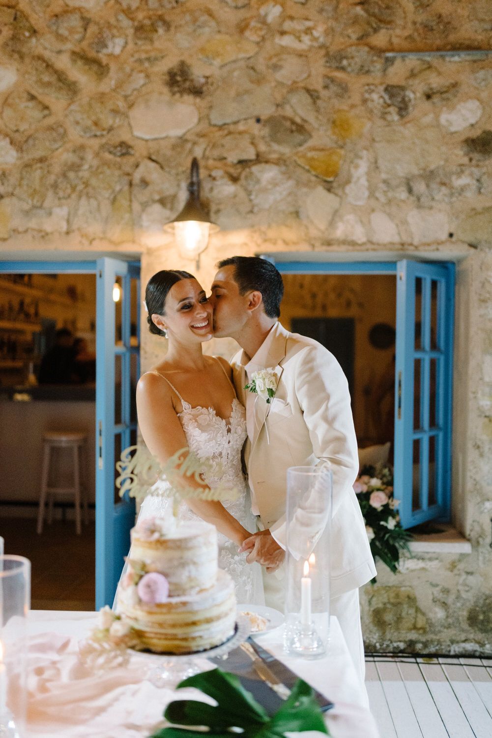 A romantic moment shared between newlyweds next to a rustic wedding cake display with stone walls and blue doors.