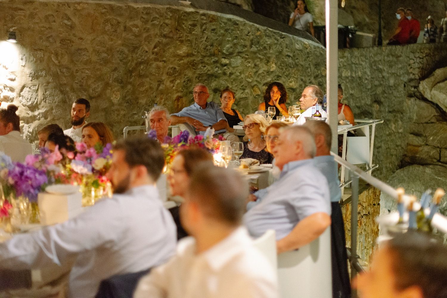 Wedding guests seated at long tables during an outdoor evening reception with stone walls and floral decor.