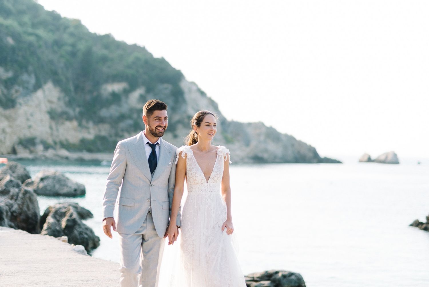 Coastal wedding portrait with couple walking on beach in light grey suit and flowing white dress.